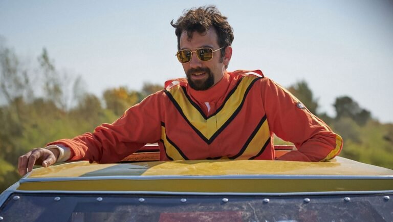 Man with a beard and sunglasses wearing an orange and yellow racing suit emerges from the roof of a yellow car.