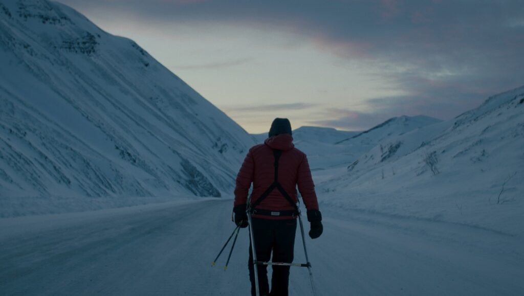 Skier in a red jacket walking away on a snowy road between steep mountains at dusk.