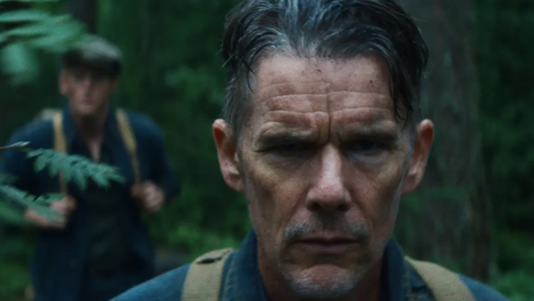 Close-up of a stern man with wet dark hair and a mustache in a forest, a blurred figure behind him.