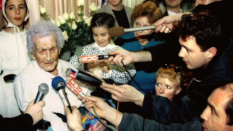 An elderly woman surrounded by journalists holding microphones labeled SUD and RMC during an interview.