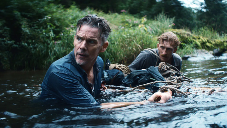 Two men wade through a shallow river while pulling a rope attached to submerged gear in a lush, green landscape.
