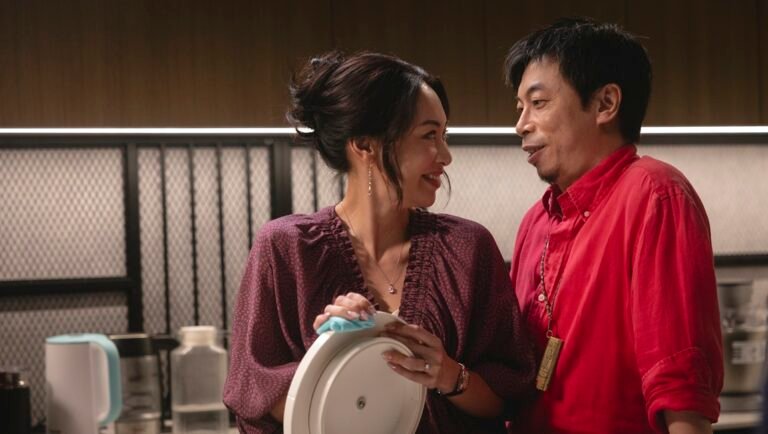 Smiling woman in a plum blouse holds a covered plate beside a man in a bright red shirt in a kitchen.