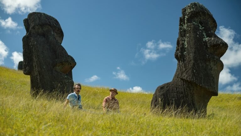 Two people sit in tall grass between two large Moai statues under a blue sky on Easter Island.