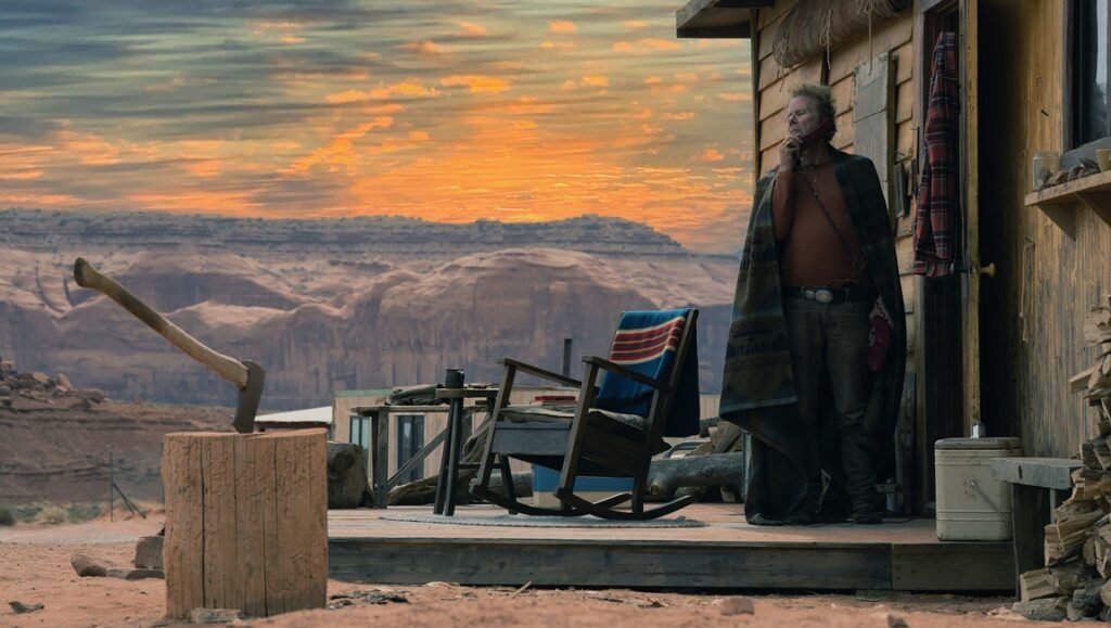 Weathered man in an open coat stands on a wooden porch, gazing at a desert sunset framed by red rock cliffs.