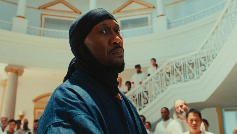 Man in a dark head wrap and blue robe looking upward inside a grand white hall with a spiral staircase and crowd.