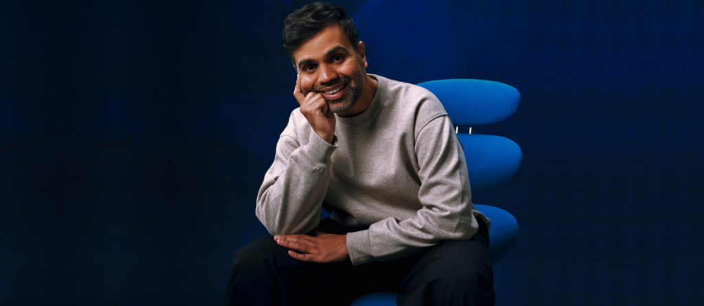 Smiling man in a gray sweatshirt resting his chin on his hand while sitting on a blue chair against a dark blue background