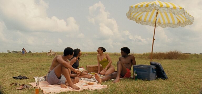 NYFF 2026 — Dispatch 2: A Long Winter, Misty Green, Clarissa Four friends sit on a blanket under a yellow striped umbrella during a picnic in a grassy field.