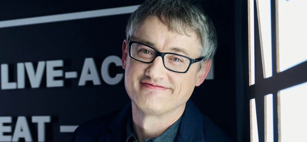 Man with short gray hair and black glasses smiling in front of a dark sign with white text.