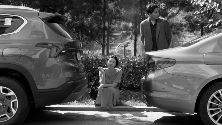Black-and-white scene of a woman crouching between two parked cars while a man in a blazer stands behind her in a wooded area.