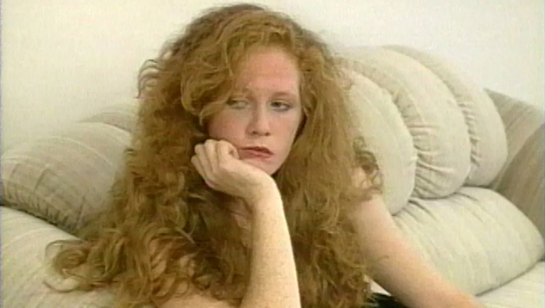 Young woman with voluminous curly red hair rests her chin on her hand while sitting on a textured beige sofa.