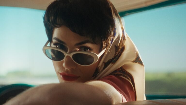 Woman with dark hair, a headscarf, and white sunglasses looking back from the passenger seat of a car.