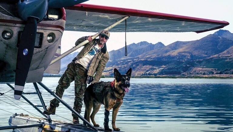 Man in camo pants and sunglasses stands on a floatplane with a German Shepherd dog on a lake.