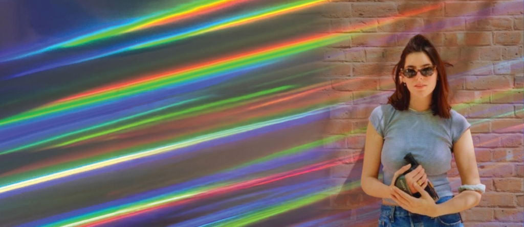 There Is No Ordinary Person: An Interview with Jacqueline Lentzou on A Day in the Life of Jo: Chapter Phaedra Woman in sunglasses and gray t-shirt holding a black object against a brick wall with diagonal rainbow light streaks.