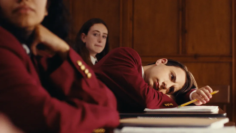 Bored student in a red blazer rests her head on a desk in a wood-paneled classroom