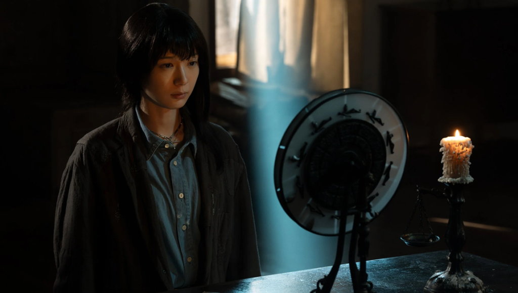 Woman with black hair standing by a candlelit desk next to a circular occult mirror in a dim room.