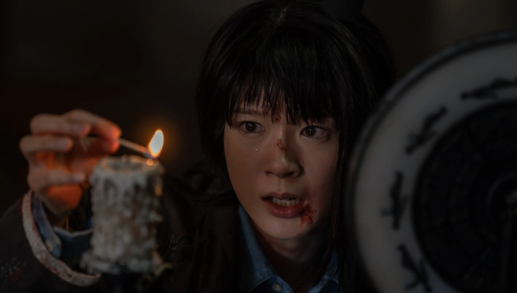 Woman with blood on her face holding a lit match over a melting candle beside a dark circular object.