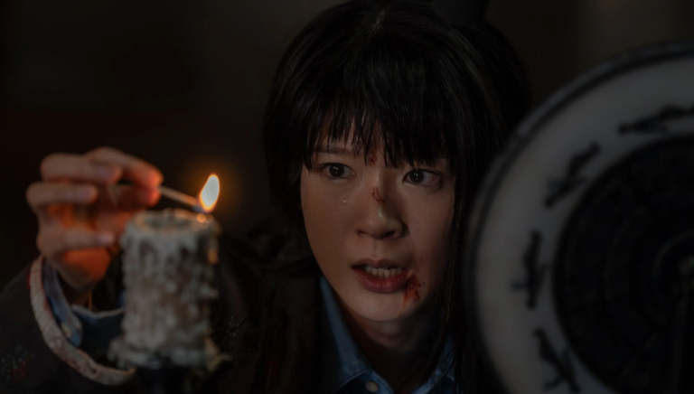 Woman with blood on her face holding a lit match over a melting candle beside a dark circular object.