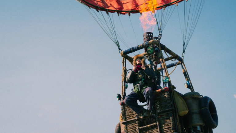 Person in a fur hat sits in a wicker hot air balloon basket as a burner shoots orange flames into the red envelope.