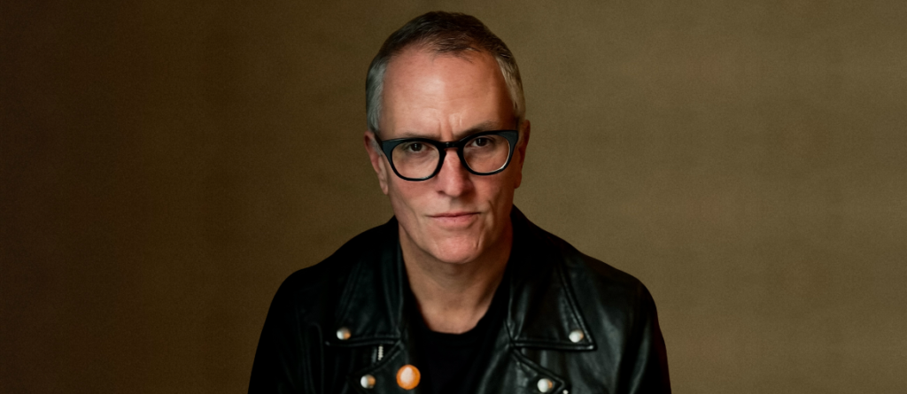 Masculinity Is Funny: An Interview With Padraic McKinley on The Weight Padraic McKinley, wearing black-rimmed glasses and a black leather jacket, poses against a brown background.
