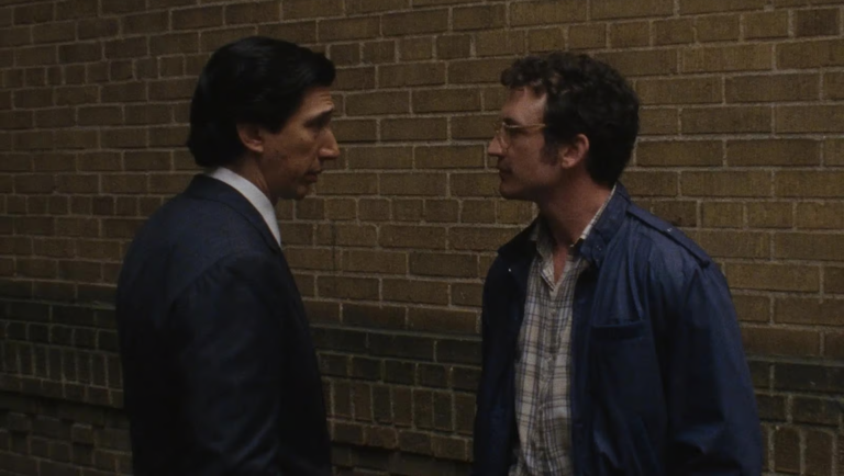Two men face each other in profile against a brick wall, one in a suit, the other in glasses and a plaid shirt.