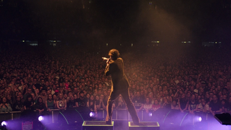 Lead singer stands on a concert stage, back to the camera, pointing toward a large, dimly lit audience.