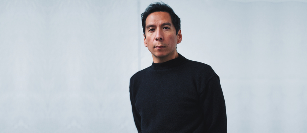 Sky Hopinka wearing a black mock-neck sweater against a light gray background.