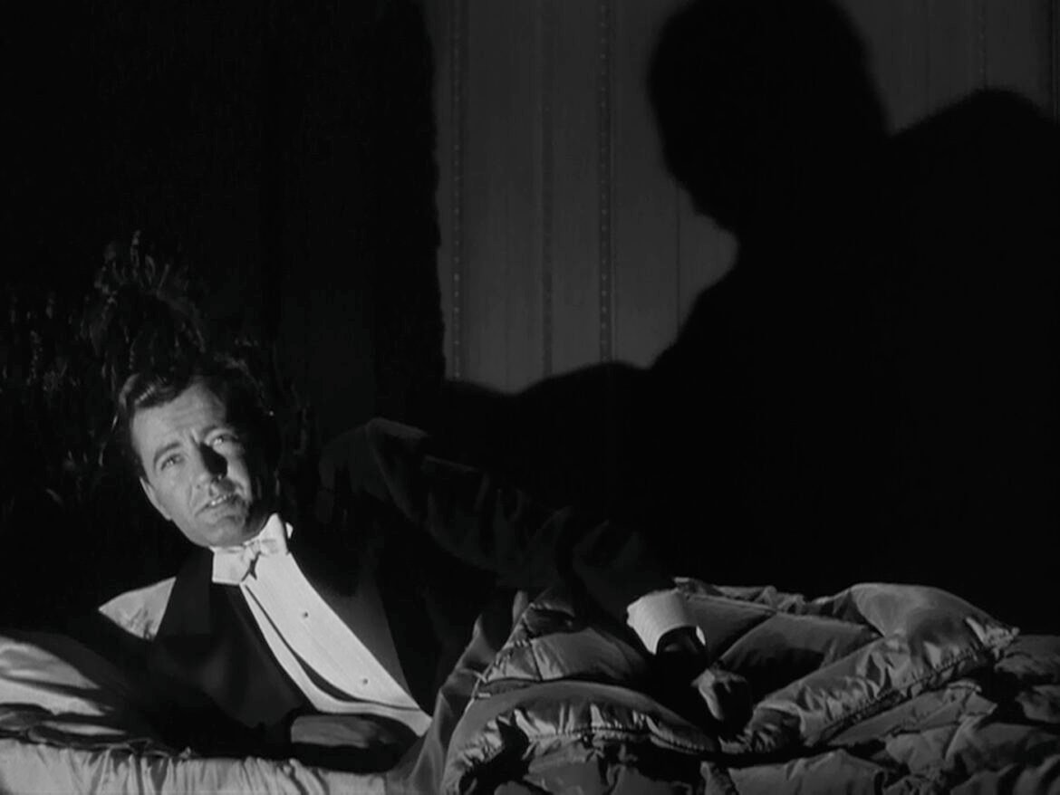 A man in a tuxedo lying in bed under low-key lighting with a large, ominous shadow cast on the wall behind him.