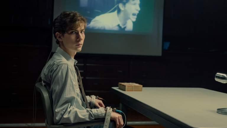 Man in a light shirt and tie sits restrained in a chair, looking back toward the camera in a dim room with a projector screen.