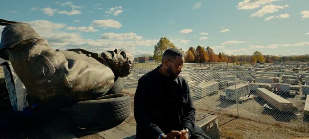 A man in a dark jacket sits beside a massive, toppled bronze head resting on a tire amid rows of concrete blocks under a blue sky.