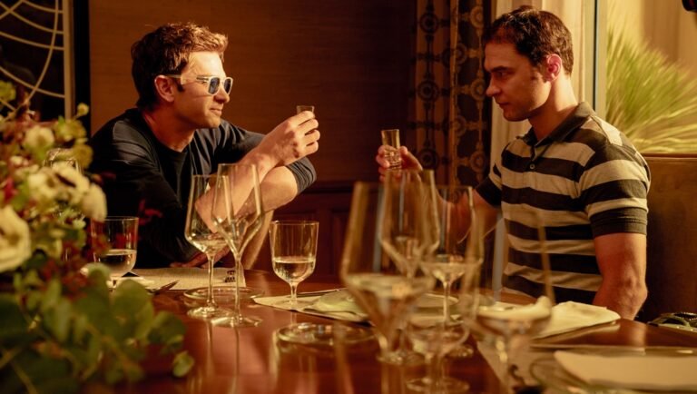 Two men sit at a candlelit table with wine glasses, one in sunglasses and a dark shirt, the other in a striped polo.