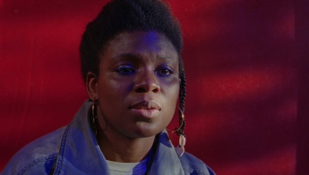 A woman with braided hair and gold hoop earrings gazes intensely against a deep crimson wall.
