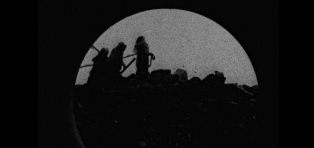 Silhouetted figures traverse a debris-laden ridge within a circular frame against a pale sky