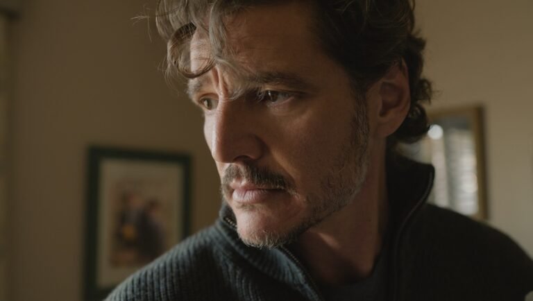 Man with a mustache and stubble looking down in a dimly lit room, wearing a dark zipped sweater.