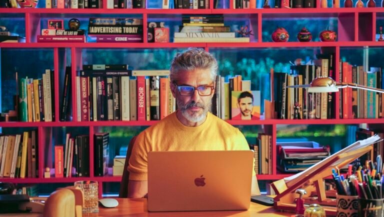 Gray-haired man in a yellow shirt typing on a laptop at a desk, backed by a red bookshelf crammed with books and art.
