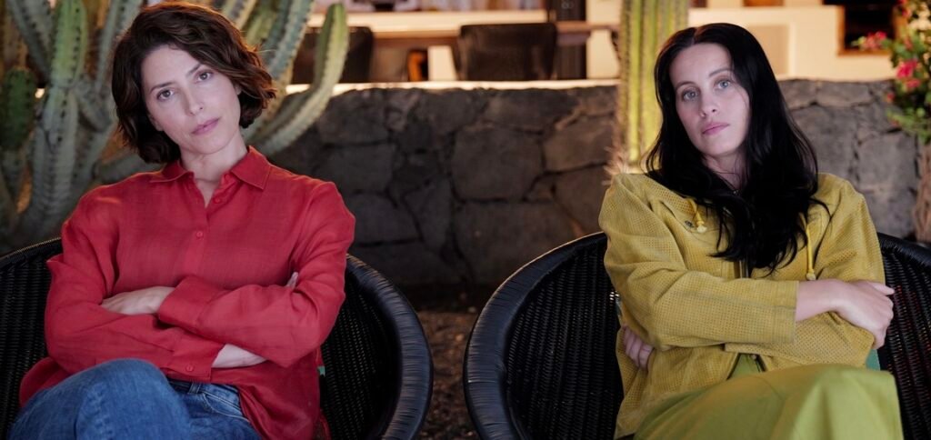 Two women sit in wicker chairs with crossed arms, one in a red shirt, the other in a yellow jacket, outdoors by a stone wall.