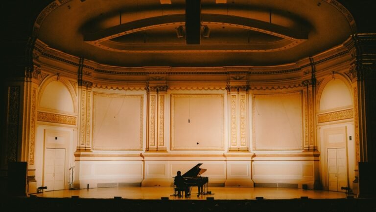 Solo pianist playing a grand piano on a warmly lit, empty concert hall stage with ornate gold walls