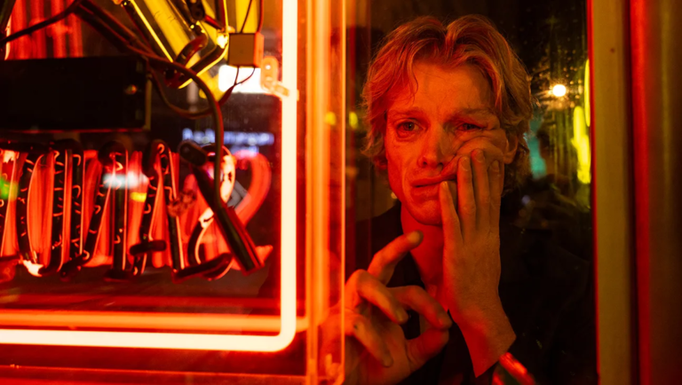 Blonde person with a hand to their face stands behind a glass window lit by red neon, with cables and glowing tubes visible inside.