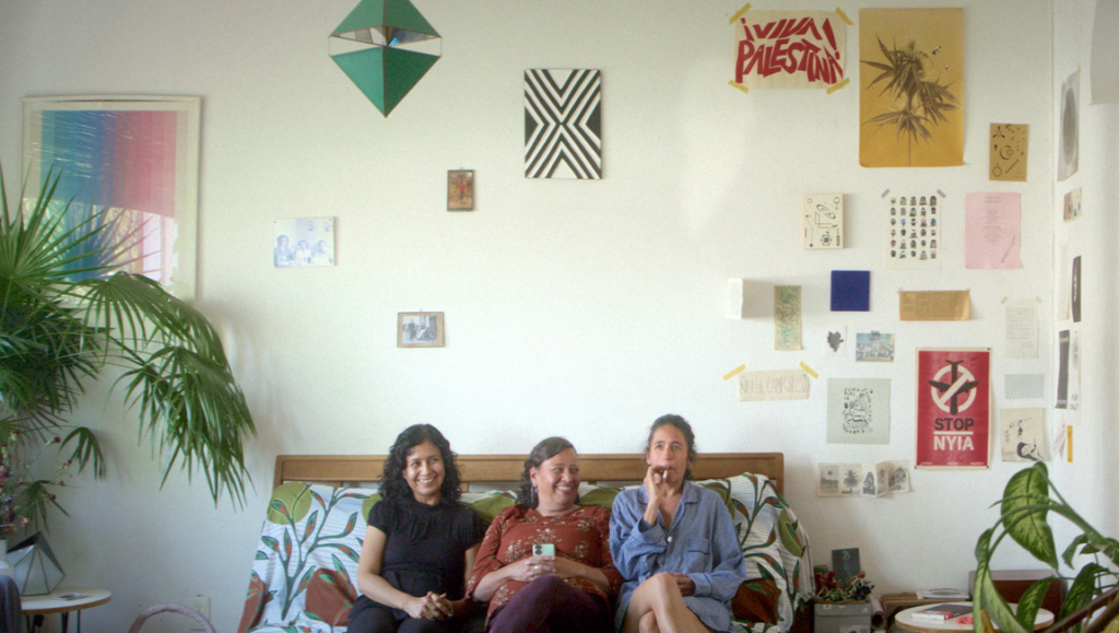 Three women sit on a patterned sofa beneath a wall covered in posters and prints, including one reading Free Palestine.
