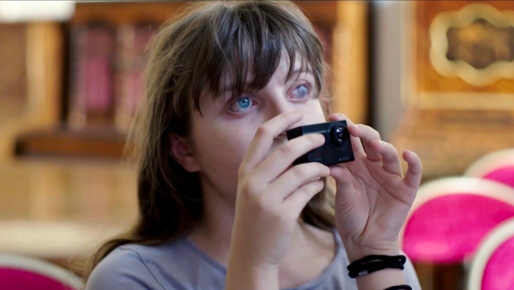 Blue-eyed girl with bangs holding a compact black camera, pink chairs blurred behind her.