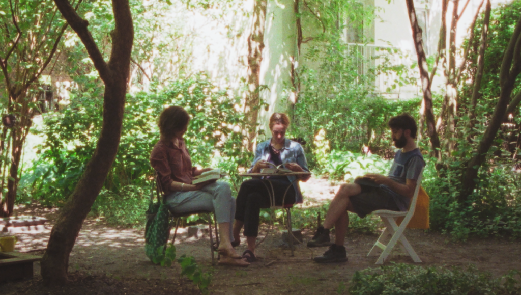 Three people reading scripts in a sunlit garden with trees and foliage.