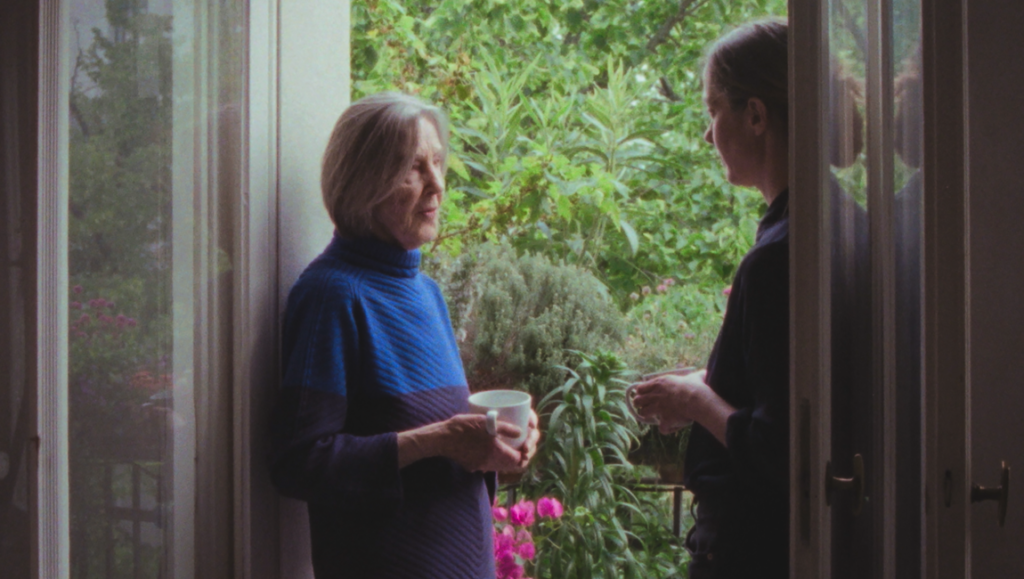 Older woman in a blue knit sweater holding a mug stands by a glass door, talking with a woman beside a lush garden.