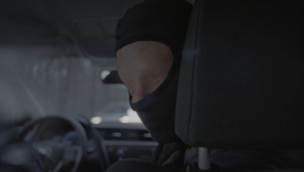 Hooded figure seen through rain-streaked glass gripping steering wheel inside dark vehicle