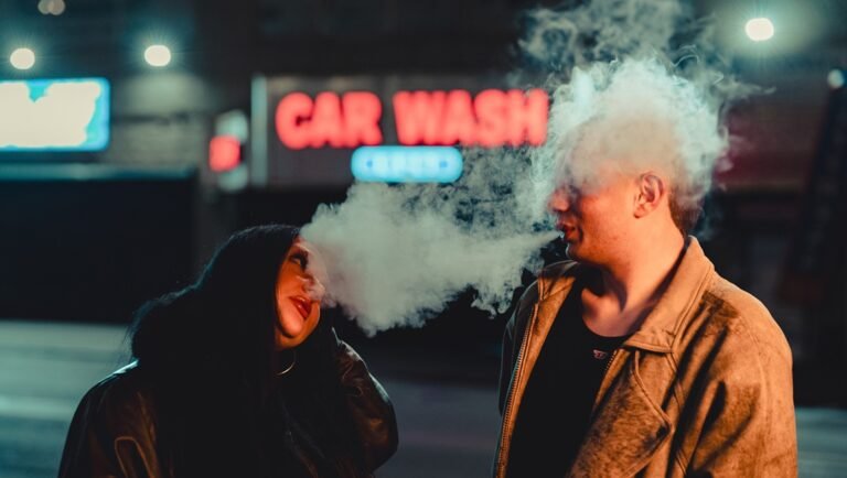 Man exhaling a thick cloud of smoke beside a woman at night in front of a neon CAR WASH sign