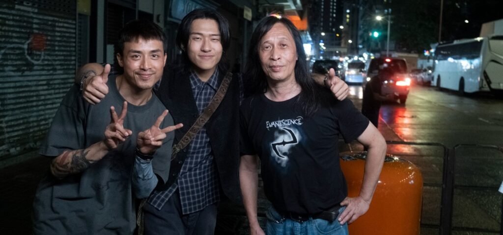 Three men pose together on a wet city street at night, one making peace signs.