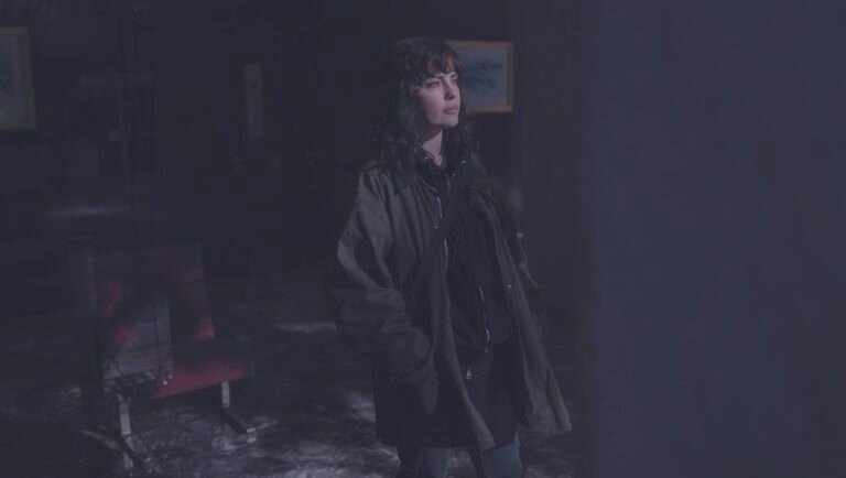 Woman in dark coat standing in a dimly lit room beside a red chair.
