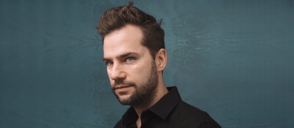 Bearded man with swept-back brown hair and blue eyes in a dark shirt against a textured teal background