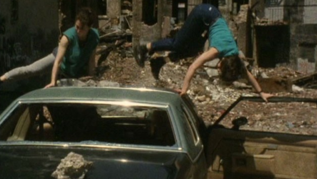 Two women in teal shirts climb over a green car amid rubble in a ruined building.