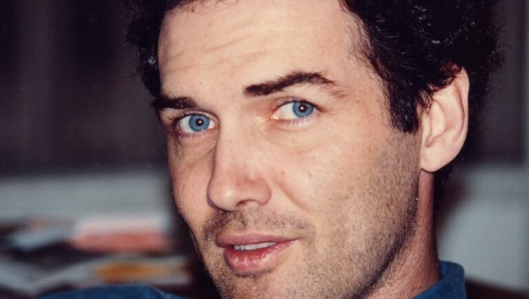"Man with blue eyes and dark curly hair, wearing a blue shirt, looking directly at the camera in an indoor setting with a blurred background."