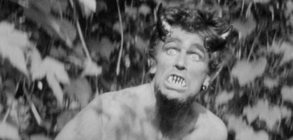 Black-and-white close-up of a person with pointed ears and fanged teeth grimacing among leaves.