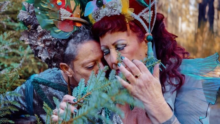Two women in ornate headdresses and makeup touch foreheads while smelling redwood branches